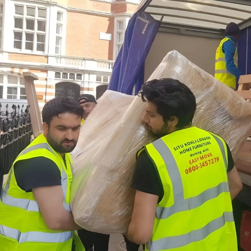 Two movers in yellow safety vests carrying a large wrapped piece of furniture into a moving truck on a city street.