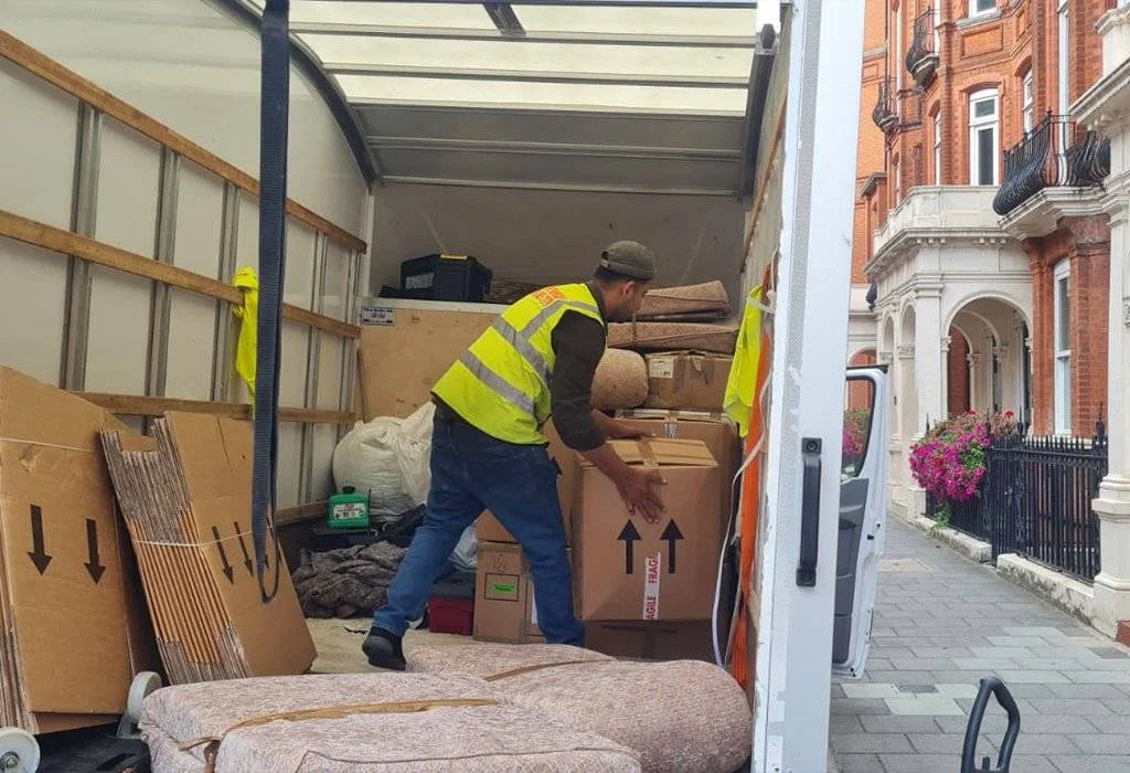 A mover in a high-visibility vest loading cardboard boxes into a moving truck parked outside residential buildings.