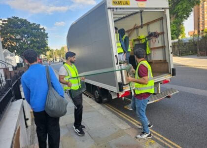 Two movers in high-visibility vests carefully carrying a glass panel toward a moving truck, with other team members organizing items inside the truck.