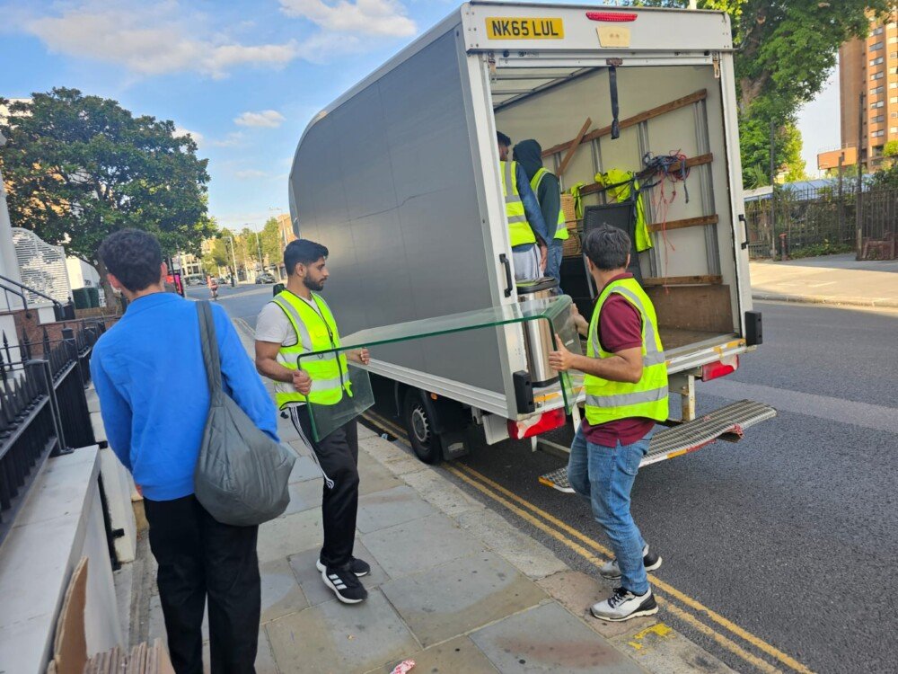 Two movers in high-visibility vests carefully carrying a glass panel toward a moving truck, with other team members organizing items inside the truck.