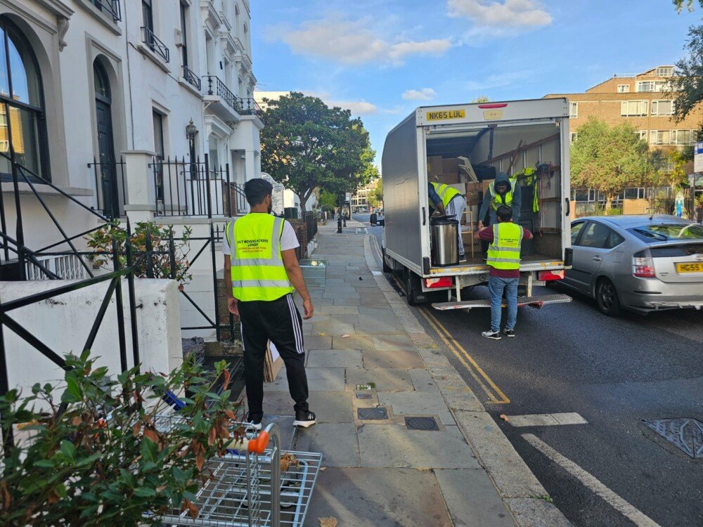 A team of movers in bright safety vests loading boxes and household items into the back of a moving truck while another mover supervises.