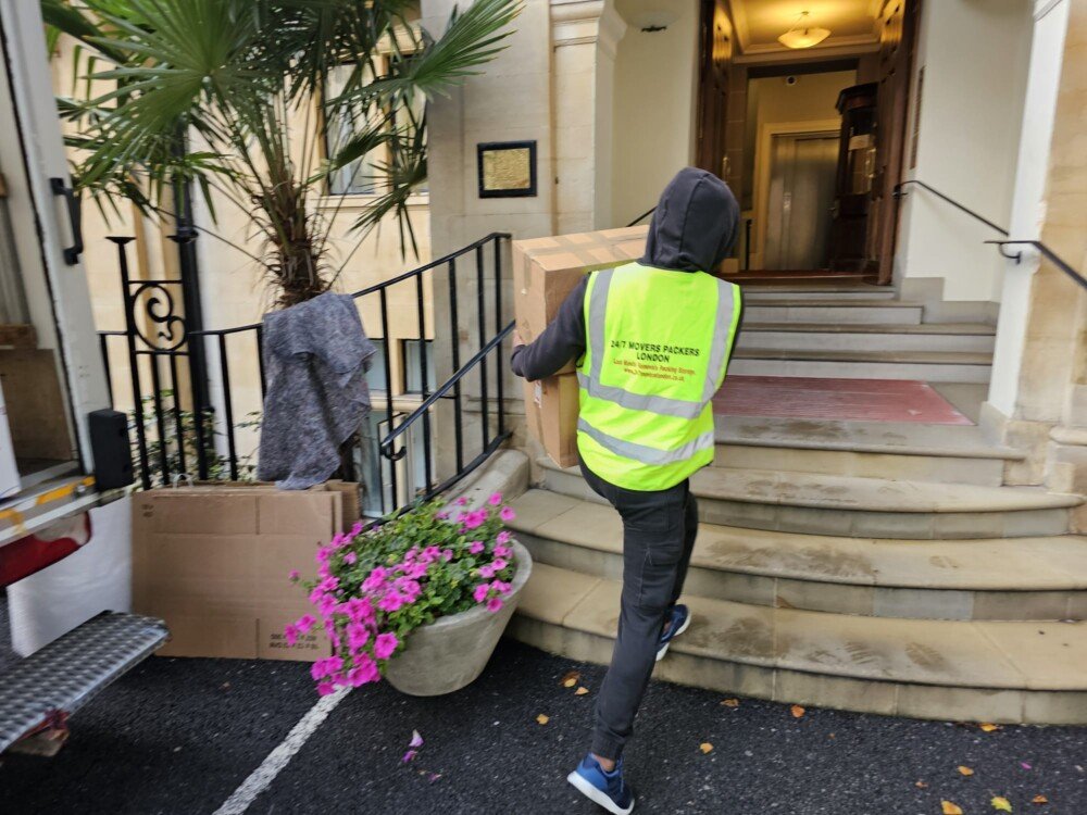 A mover in a fluorescent vest carrying a cardboard box up the steps into a building entrance.