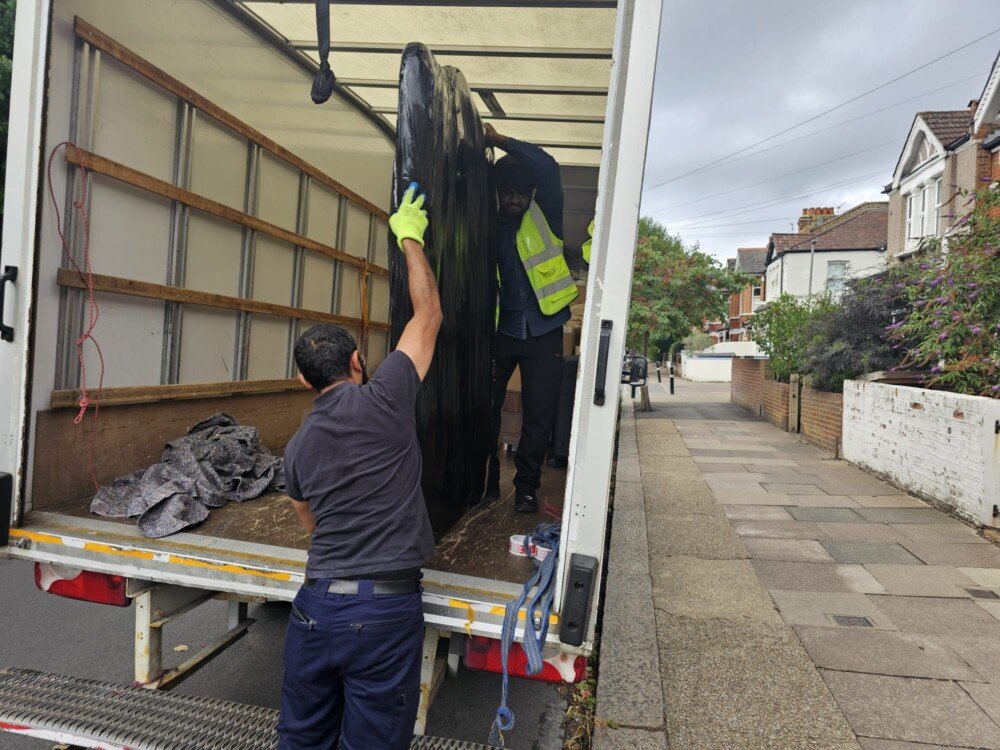 Two movers loading a large black item into the back of a moving truck parked on a residential street.